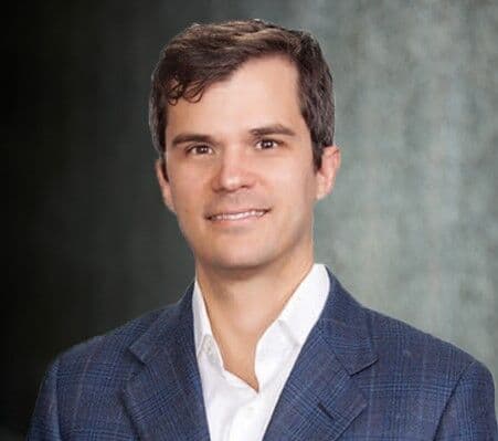 Head-and-shoulders portrait of a smiling Dr Luke Tomycz in a blue suit jacket and white shirt against a gray textured background.