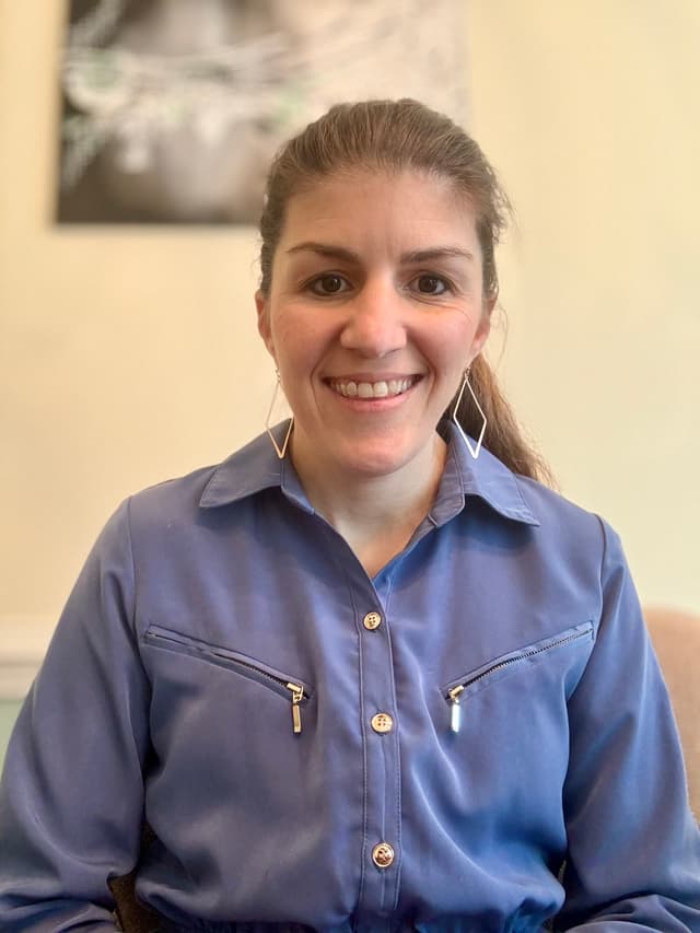 Close-up smiling Taisa Meighan with long brown hair wearing a navy-and-white striped top, seated indoors.