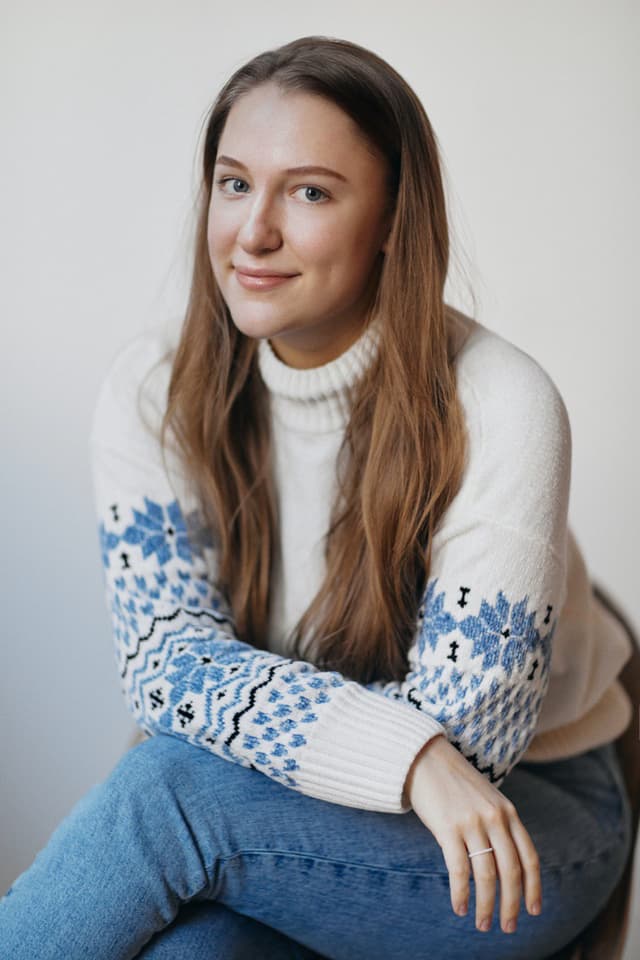 Head-and-shoulders portrait of a smiling Yulia Aliabieva with long brown hair wearing a white turtleneck sweater with blue patterned sleeves, seated against a neutral background.
