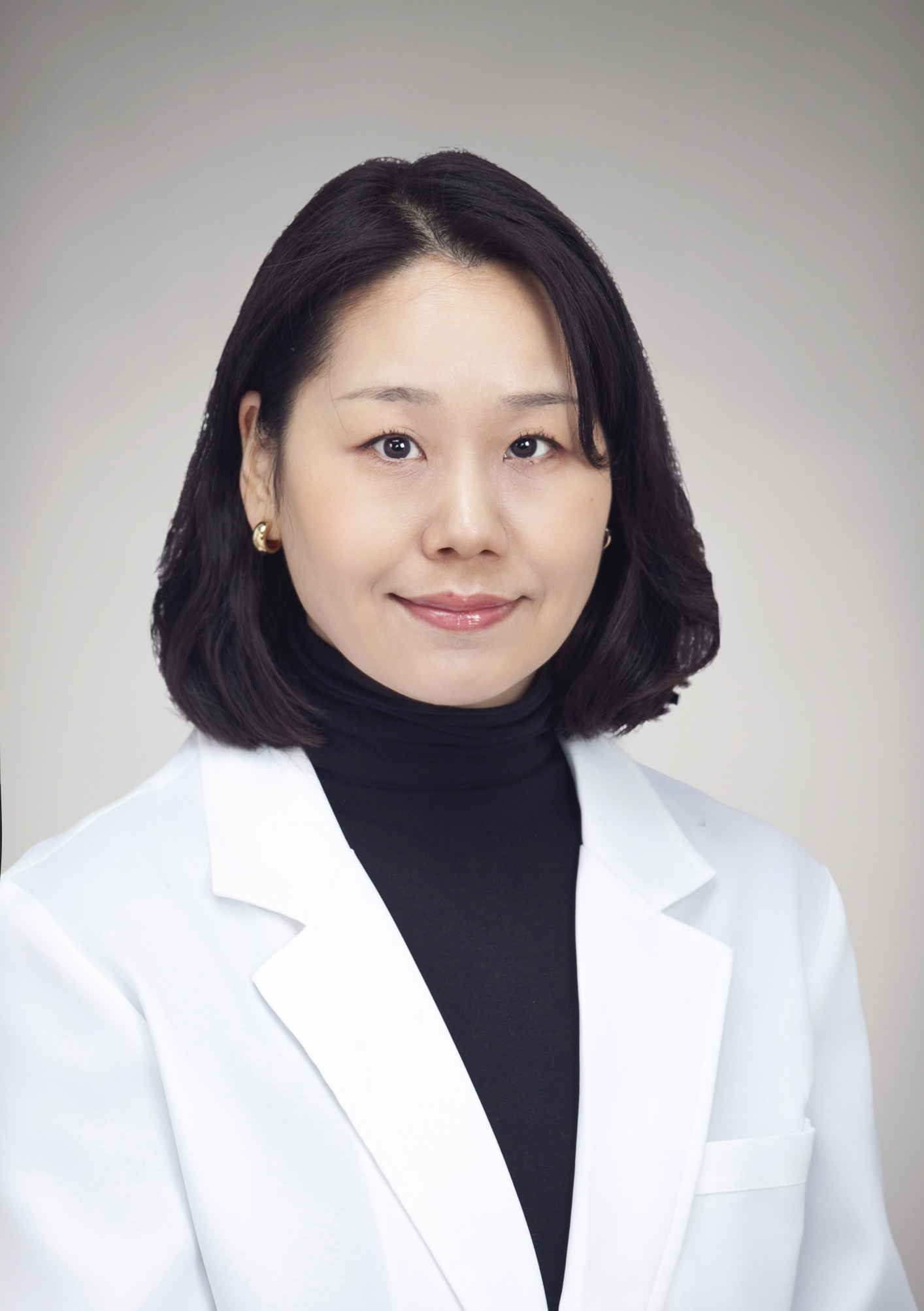 Head-and-shoulders portrait of a smiling Dr Alma Yum in a white coat and black turtleneck against a neutral gray background.