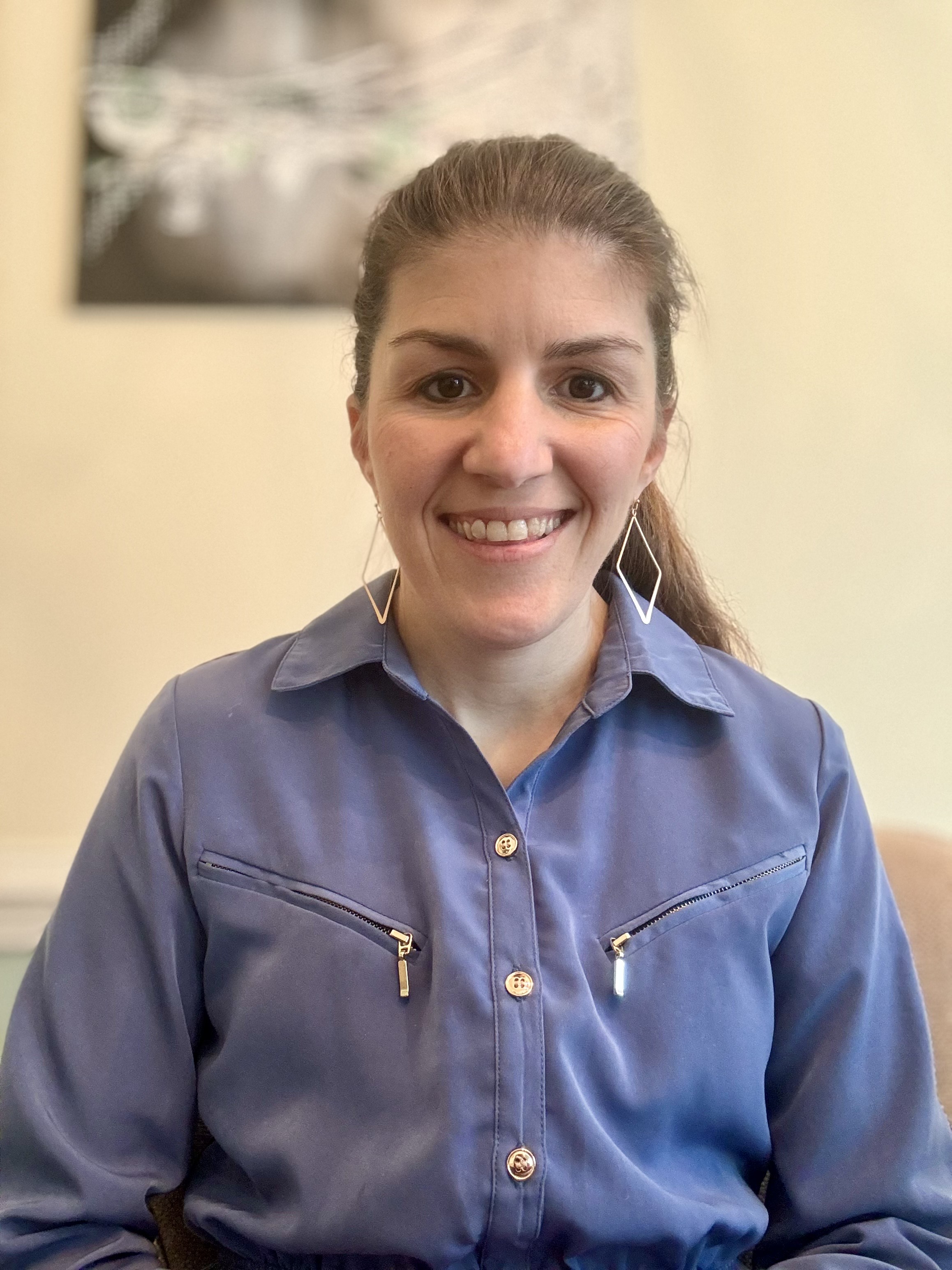 Close-up smiling Taisa Meighan with long brown hair wearing a navy-and-white striped top, seated indoors.