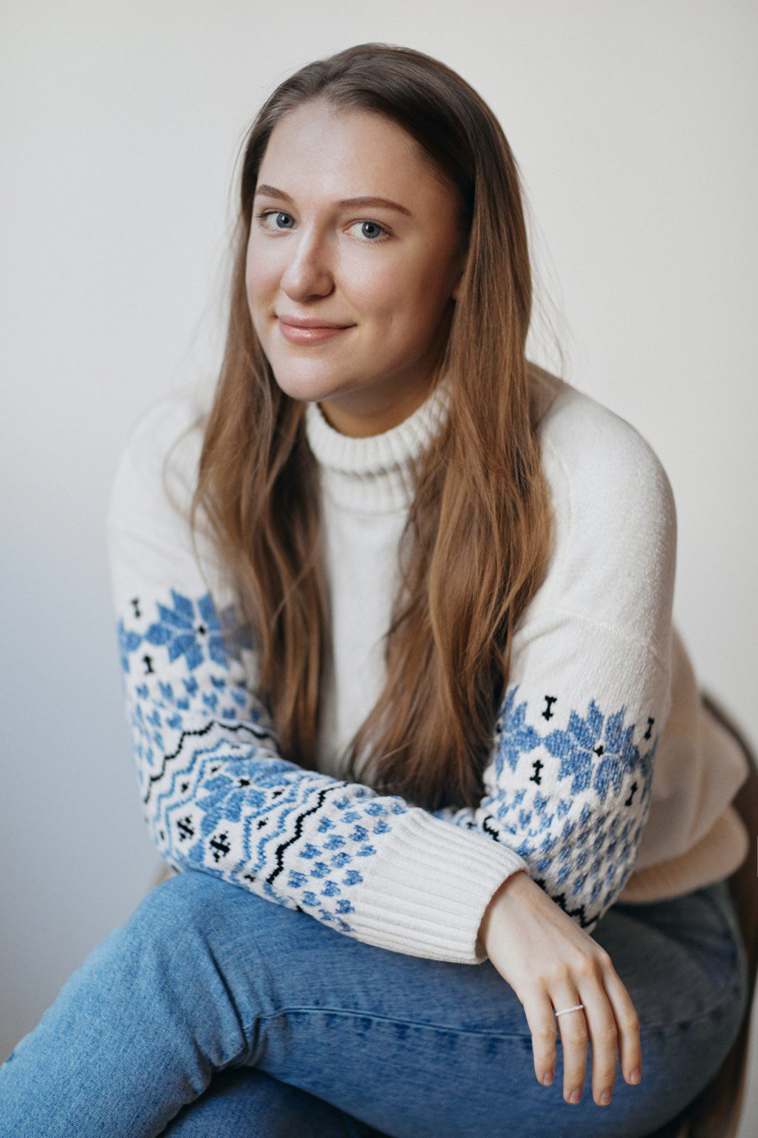 Head-and-shoulders portrait of a smiling Yulia Aliabieva with long brown hair wearing a white turtleneck sweater with blue patterned sleeves, seated against a neutral background.